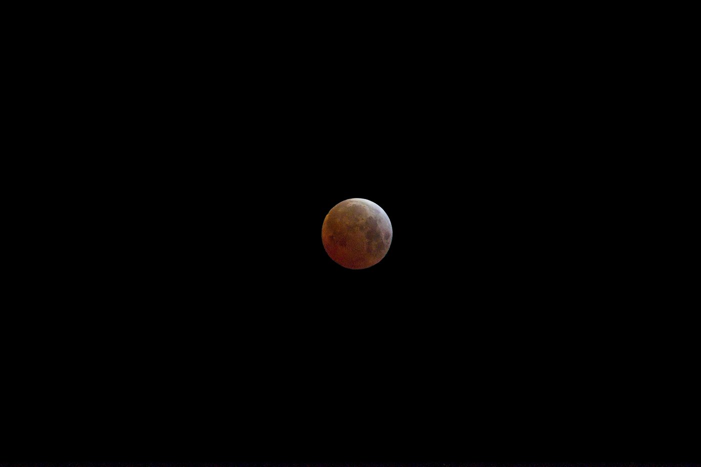 A blood moon captured during a total lunar eclipse, glowing with a reddish-orange hue against a pitch-black night sky. The Moon's surface displays subtle craters and maria patterns, with a slightly darker shadowed portion visible along one edge, indicating the Earth's shadow partially covering the lunar disk.